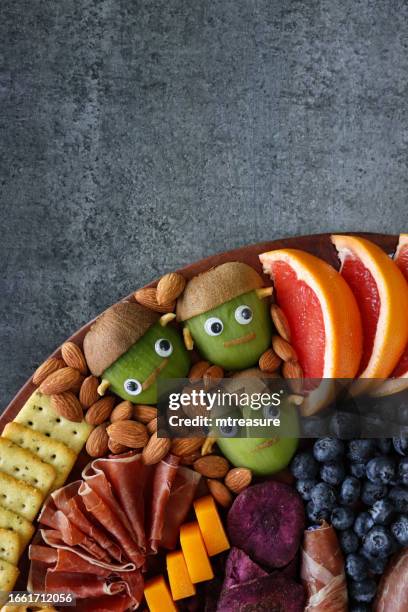 close-up image of cheese-charcuterie board, frankenstein monster kiwi fruit, prosciutto di parma, pink grapefruit, almonds, beetroot crisps, cheese cubes, crackers, blueberries, grey background, elevated view, copy space - frankenstein stock pictures, royalty-free photos & images