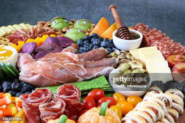 close-up image of halloween cheese-charcuterie board, prosciutto di parma hand, pumpkin brie, honey dipper and ramekin of honey, cucumber, frankenstein monster kiwis, salami roses, beetroot crisps, blueberries, grey background, focus on foreground - frankenstein stock pictures, royalty-free photos & images