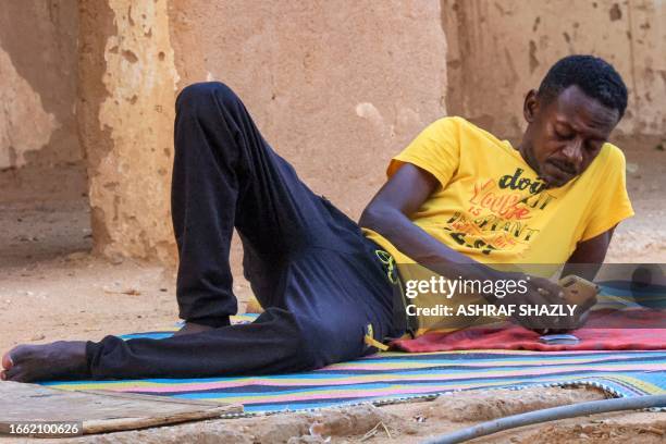 Man browses a phone while lying next to playing cards on a straw mat outside a school that has been transformed into a shelter for people displaced...