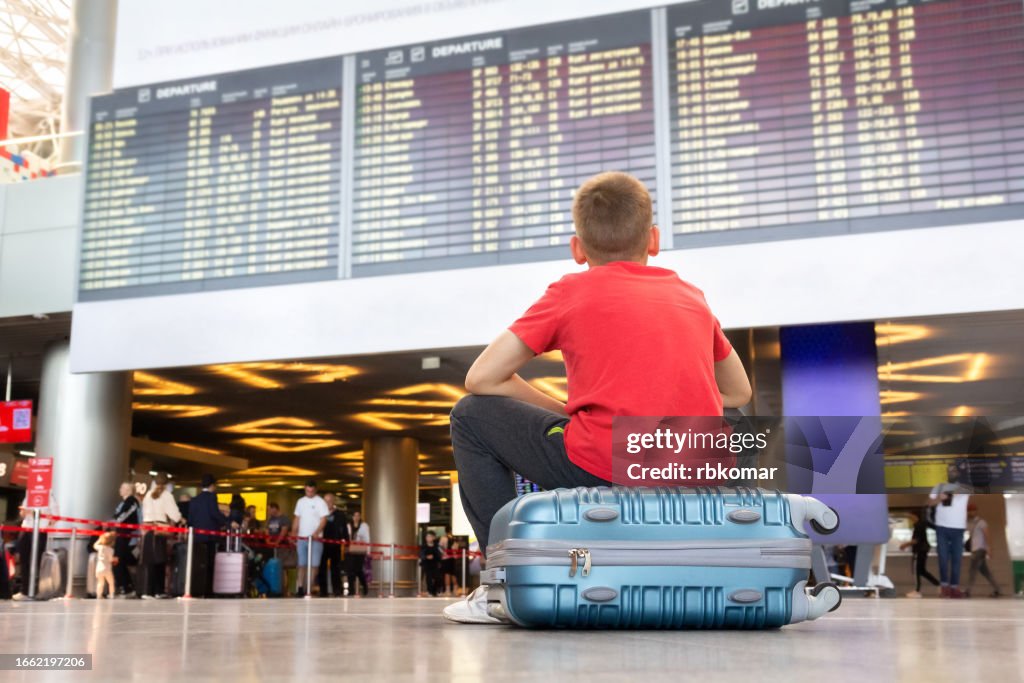 Lonely child sitting on a suitcase and waiting for departure at the airport looking at the arrivals board