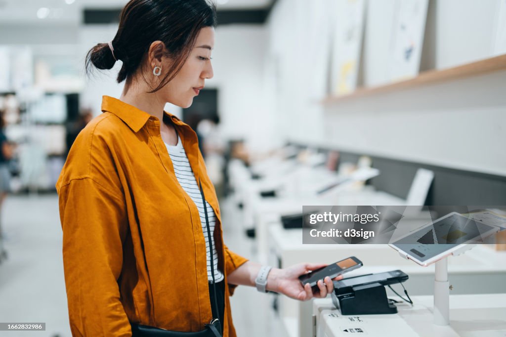 Young Asian woman using contactless payment via smartphone to pay for her shopping at self-checkout machines in a store. Innovation and technology