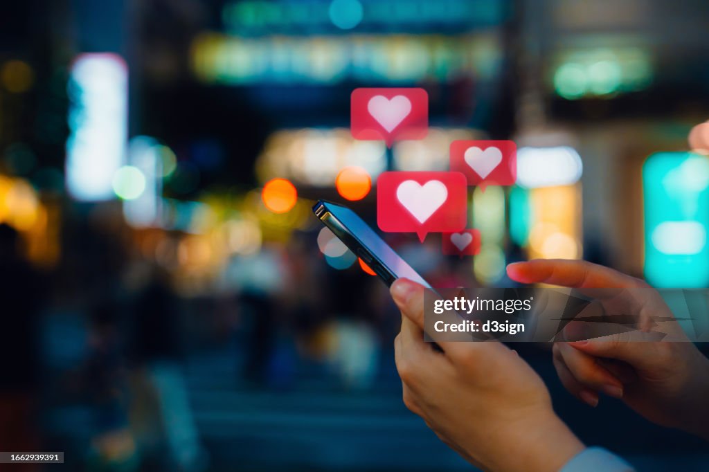 Young woman connecting with social media network on smartphone. Receiving notifications, viewing posts, giving likes on social media posts. With heart shape emoji speech bubble message on device screen. People engaging in networking with technology
