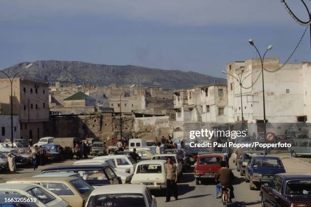 Pedestrians pass cars parked on a square in the Medina of Fez within the city of Fes in the Atlas Mountains region of Morocco in North Africa in...