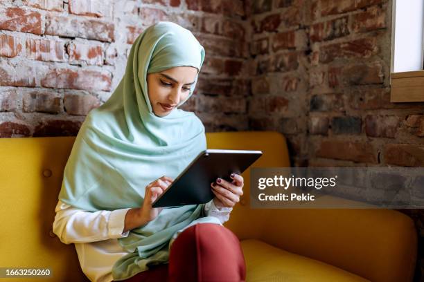 young beautiful businesswoman wearing hijab using digital tablet while sitting on a sofa at modern office - hijab stock pictures, royalty-free photos & images