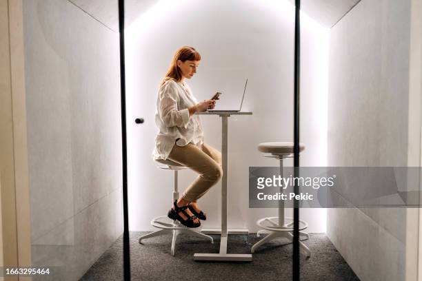 young pregnant businesswoman using phone while working in an office pod - booth stock pictures, royalty-free photos & images