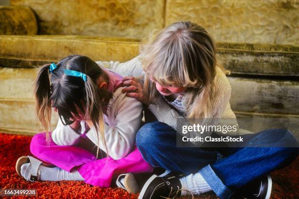 1980s Two little girls sisters sitting side-by-side on floor by sofa one girl comforts the other crying holding head in hands.