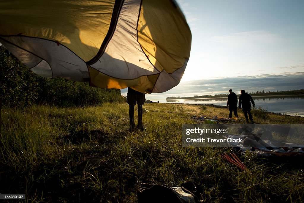 Boys taking down a tent by a river in golden morning light.
