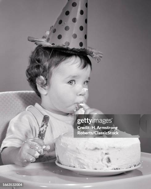 1960s Messy brunette baby with birthday cake wearing party hat icing all over mouth nose hands.