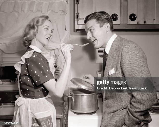 1950s Woman housewife in print dress and apron holding tasting spoon to man husband's mouth in kitchen.