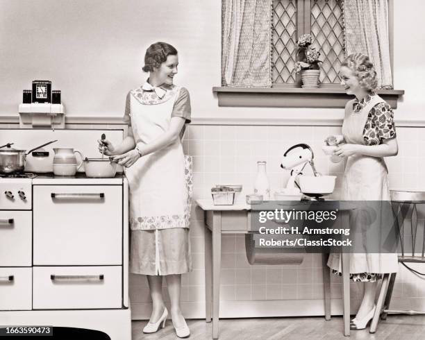 1930s Two women talking smiling while in the kitchen one stirring pot on stove other adding ingredients to electric mixer.