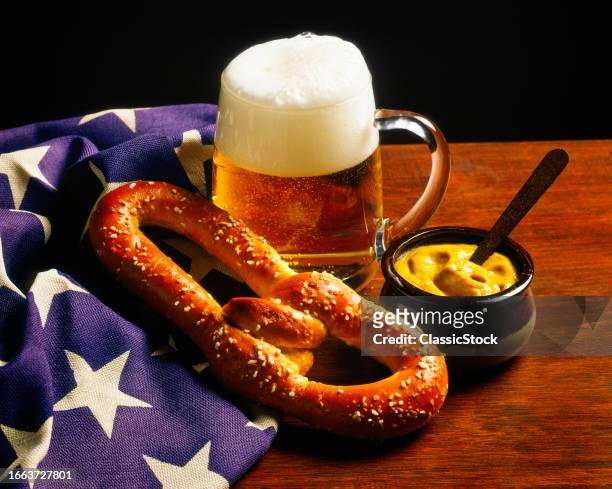 1980s Mug of beer salty soft pretzel jar of mustard with american flag on table top symbolic still life.