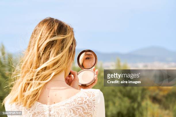 woman standing with her back looking at her reflection in a compact powder in nature outside the city - compact mirror stockfoto's en -beelden