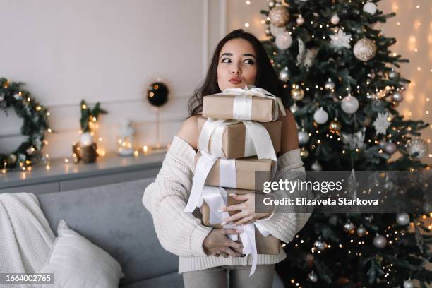 portrait of a young asian woman in a knitted sweater holding a lot of christmas gift boxes and smiling looking at the camera in a decorated room with a christmas tree in december in the apartment. - december stockfoto's en -beelden