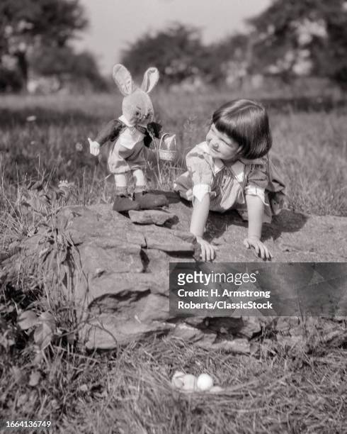 1920s Little girl leaning on rocks with stuffed easter bunny toy peter rabbit beside her looking down on nest of easter eggs.