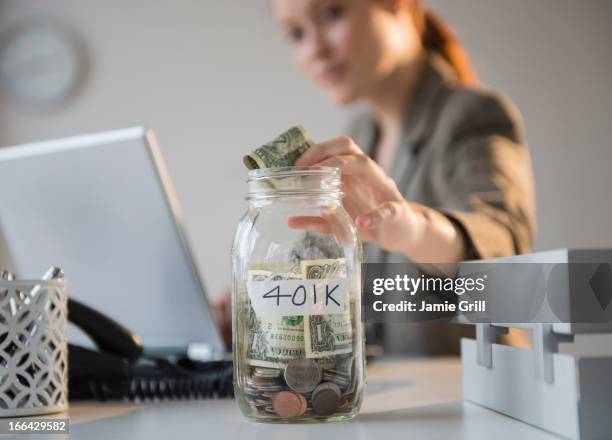 businesswoman putting money into 401k jar at desk - 401k stock pictures, royalty-free photos & images