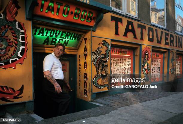 Denmark. Danemark, Copenhague- juin 1967- un homme pose debout, tatouage sur le bras, devant une boutqiue de tatouage.