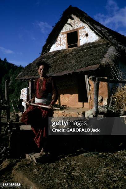 Nepal. Népal- mars 1970- Une paysanne portant une bêche, devant sa maison au toit de chaume et au fronton décoré de motifs sur fond blanc.