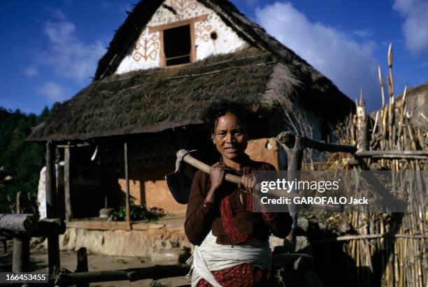 Nepal. Népal- mars 1970- Une paysanne portant une bêche, devant sa maison au toit de chaume et au fronton décoré de motifs sur fond blanc.