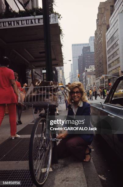 New York, United States. New York - août 1971 - Dans une rue, une femme souriante accroupie sur un trottoir, manipulant le cadenas de son vélo...