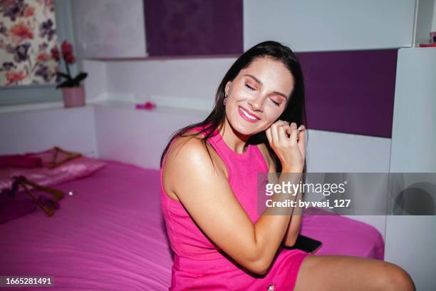 a beautiful gen z girl, eyes closed smiling, wearing hot pink clothes,  sitting on the bed in her bedroom, taking off her earrings, on camera flash effect - pink dress stock pictures, royalty-free photos & images