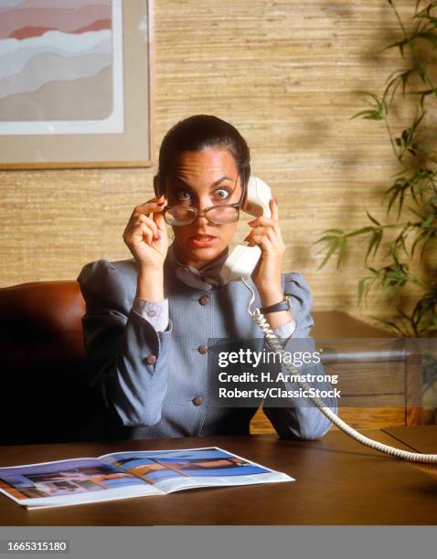 1980s Business woman on telephone call sitting at desk shocked facial expression looking over top of eyeglasses eyes wide open.