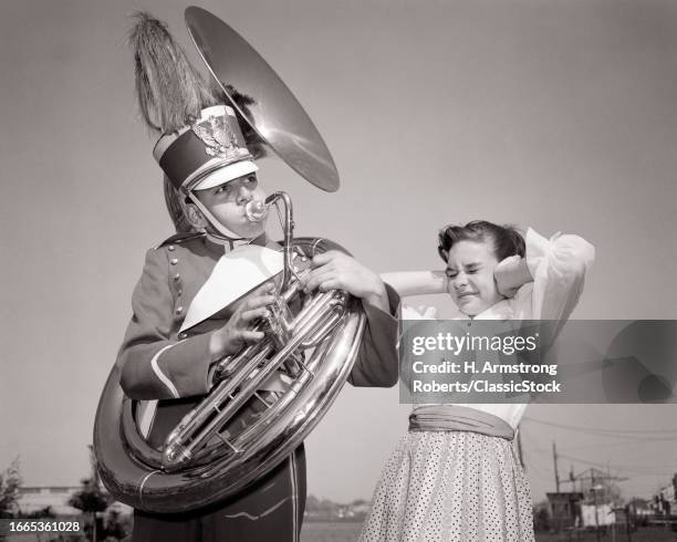 1950s Teen boy playing tuba loudly wearing band uniform and hat young girl holding her hands over her ears from the noise.