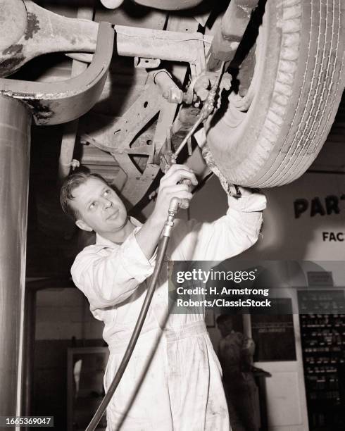 1950s Man garage mechanic wearing white coveralls working lubricating rear suspension on car on a hydraulic lift.