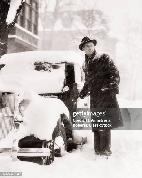 1930s Man standing next to parked car covered in ankle deep winter snow.