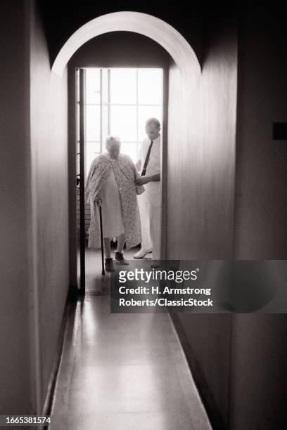 1960s 1970s male orderly helping a senior woman patient with a cane walking along an arched corridor.