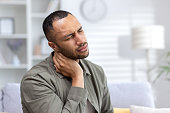 Tired young African American man sitting at home and holding his neck with eyes closed, suffering from pain and overtiredness. Close-up photo