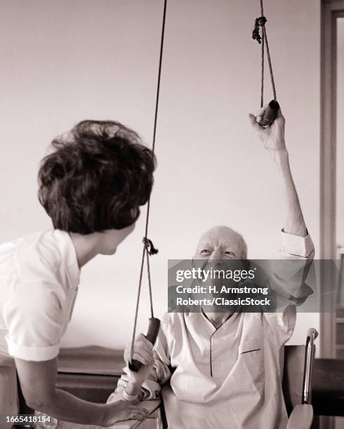 1960s Nurse physical therapist helping a senior man with pulley exercises to rehab his upper body and shoulder strength.