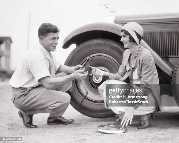 1920s Smiling couple man and woman working together to change wheel with flat tire on automobile.