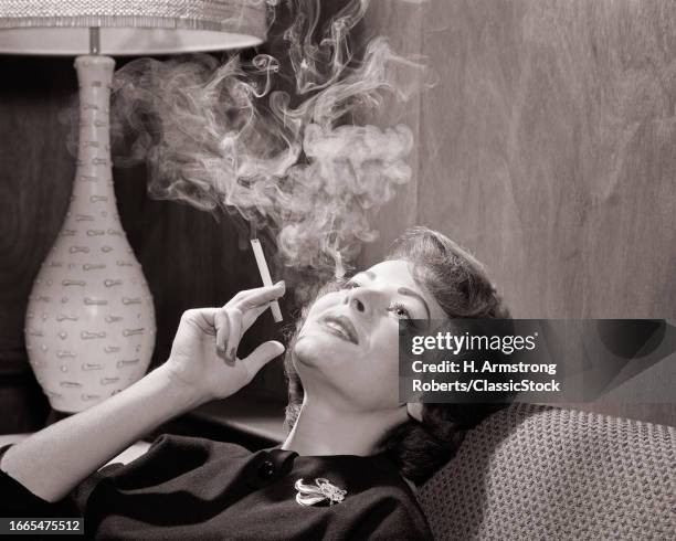 1950s 1960s relaxed pensive woman leaning back on couch enjoying smoking a cigarette smoke curling in the air above her head.