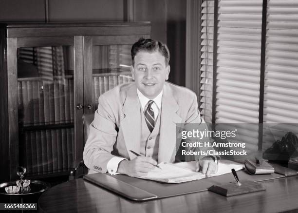 1950s Smiling business man looking at camera wearing three piece suit sitting at desk signing papers.
