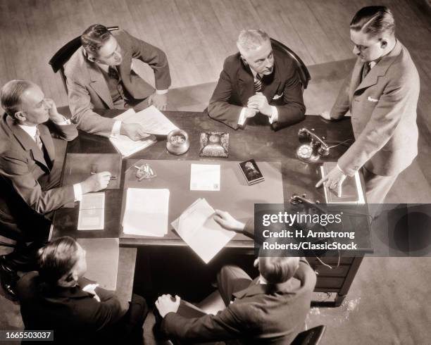 1930s Six men one standing speaking business boardroom meeting conference papers telephones and ashtrays on the table desk.