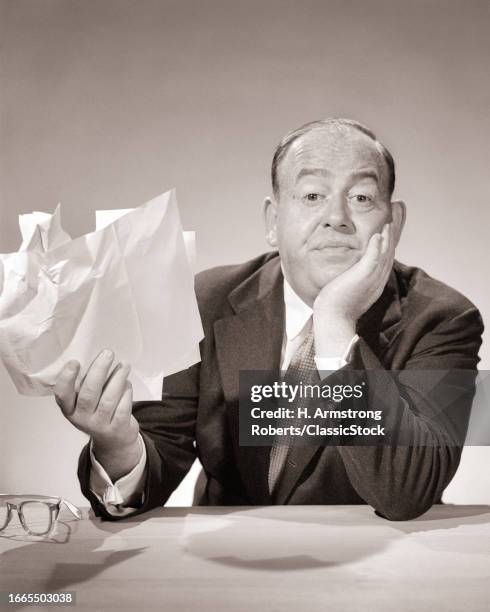 1950s Annoyed balding businessman with handful of papers resting his chin in other hand looking at camera with fed up expression.