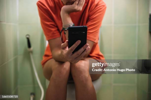 woman sitting on a toilet holding a mobile phone in her hands - woman hemorrhoids fotografías e imágenes de stock