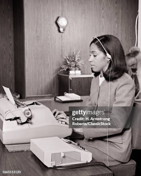 1970s woman in office typing on electric typewriter listening to dictation from dictaphone with headset.