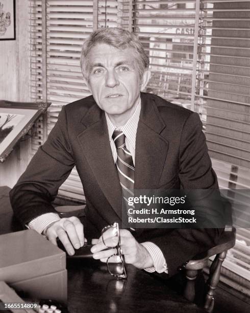 1970s Mature man businessman in suit and tie holding glasses sitting behind office desk.