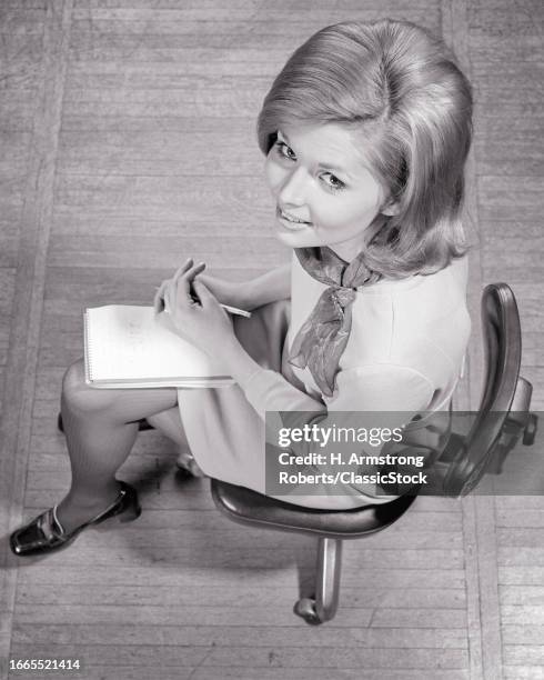 1960s Woman secretary stenographer sitting in office chair steno pad & pencil on lap looking up at camera.