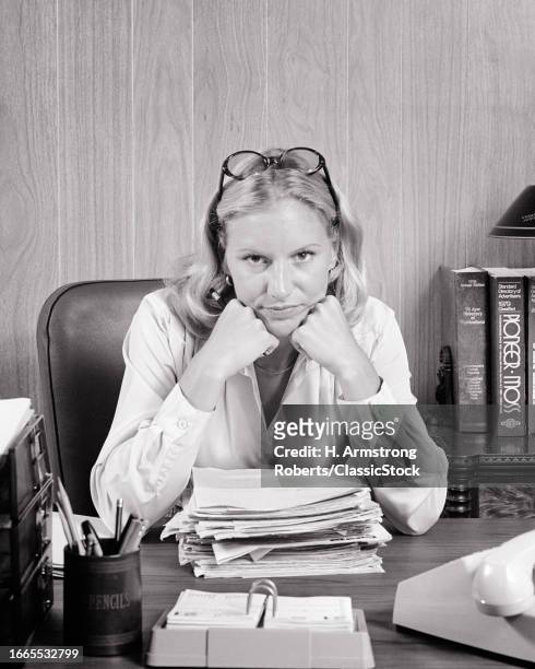 1970s Disgruntled overworked blonde woman leaning face on her hands elbows on desk above a pile of paperwork looking at camera.