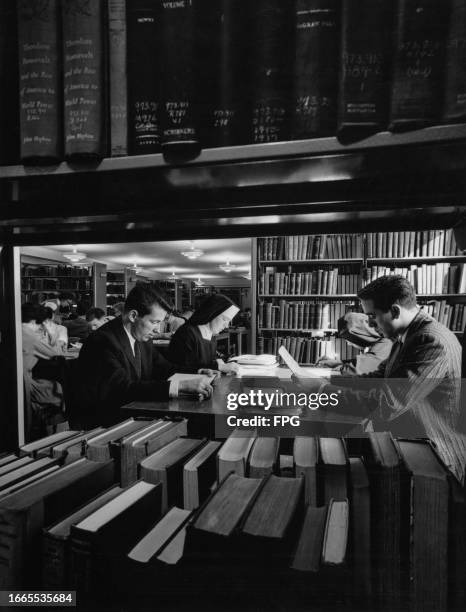 Group of people, including two nuns, sit around a table on which are books, in a reference library, United States, circa 1955.