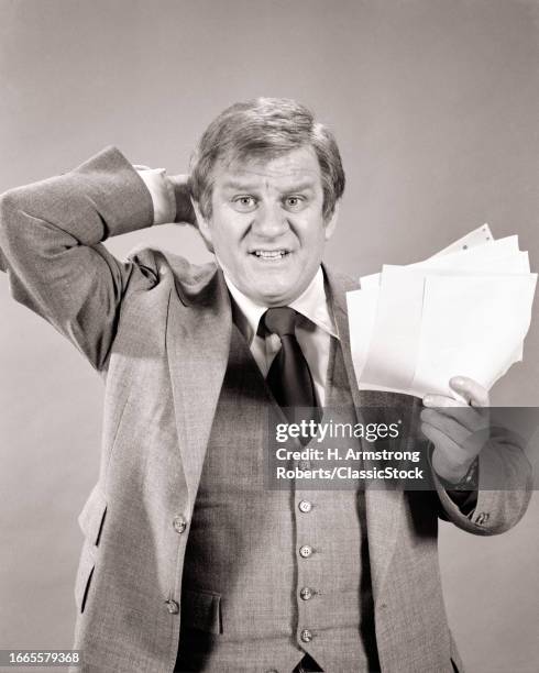 1970s Concerned businessman holding papers bills hand behind head looking at camera.