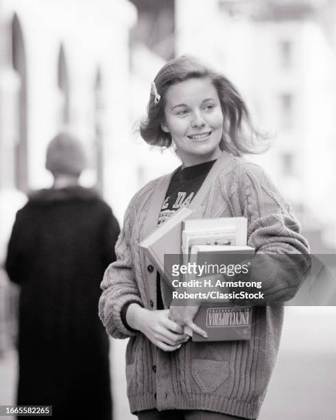 1960s 1970s smiling coed college girl holding an armful of books textbooks wearing bulky sweater looking to side.