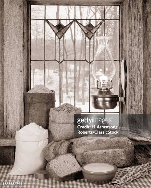 1970s Bags of grains flour slices loaf bread bowl butter in front of stained glass window hurricane lamp winter scene outside.