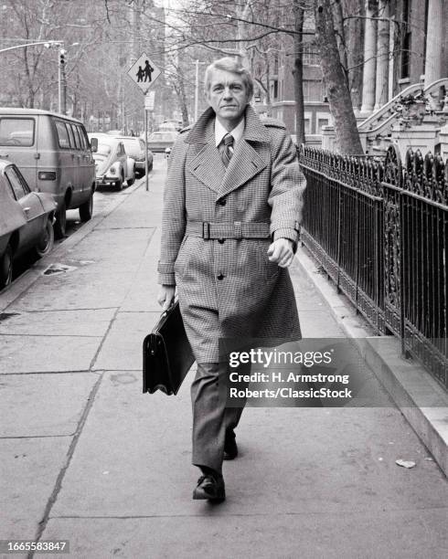 1970s Unsmiling middle-aged man businessman salesman wearing belted wool coat walking down city street carrying briefcase.