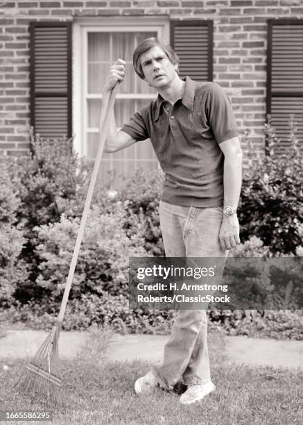 1970s Man standing in front of brick house leaning on rake tired lazy facial expression.