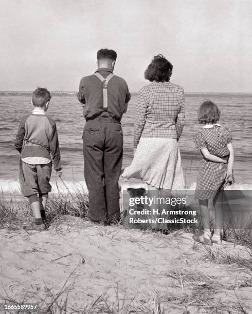 1930s Rear view american family with dog on seashore beach sand dune looking out at atlantic ocean barnegat city new jersey USA.