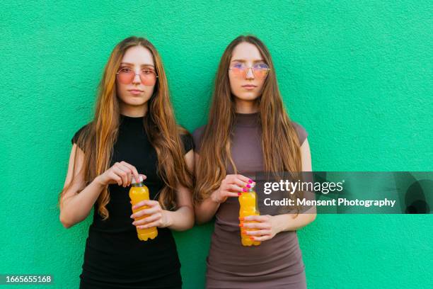 young gen-z twin sisters with cool sunglasses holding yellow lemonade plastic bottles standing on a green wall background - cute girlfriends stock pictures, royalty-free photos & images