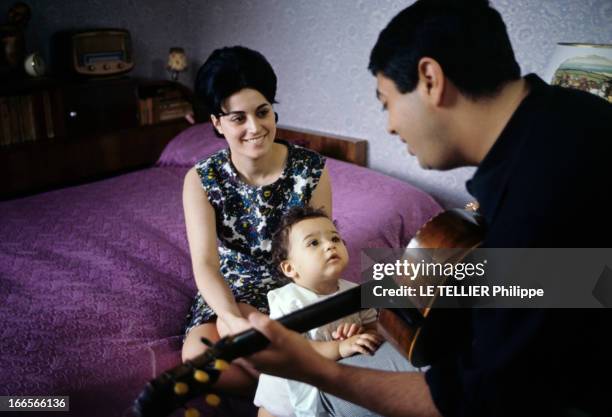 Close-Up Of Enrico Macias. En 1964, Enrico MACIAS, de dos, jouant de la guitare, en compagnie de sa femme Suzy, assise sur un lit, tenant leur fille...